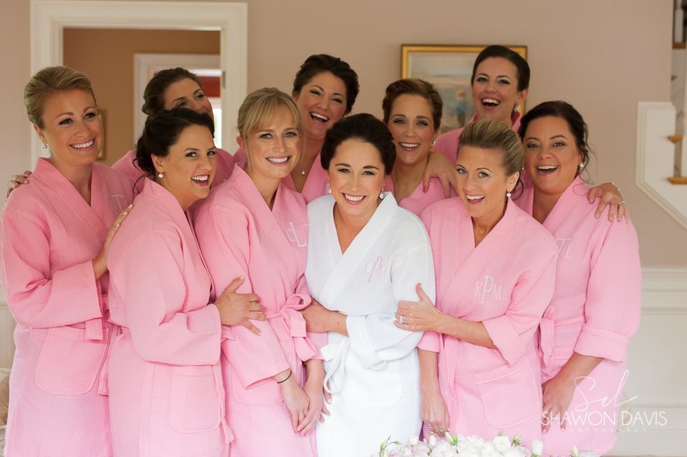 Women in pink robes surround a woman in a white robe, smiling indoors before a wedding