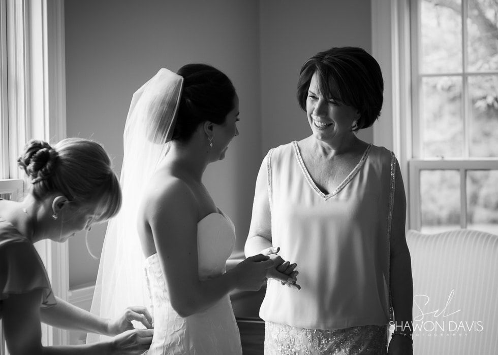 Bride and woman smiling as they stand indoors, while another woman adjusts the bride’s dress.