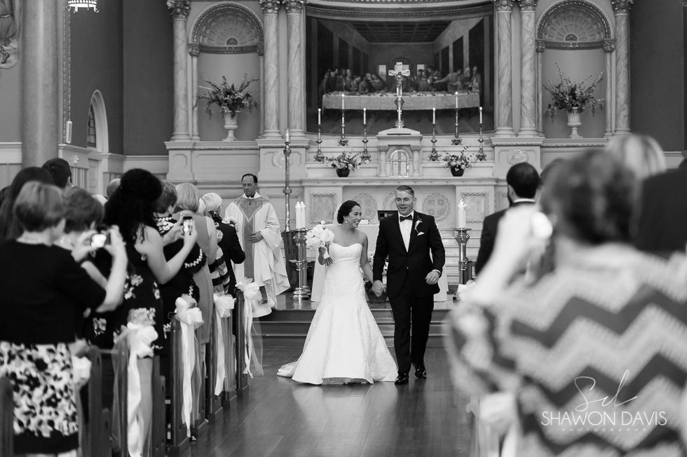 Bride and groom walk down a church aisle during a wedding ceremony, guests applauding.