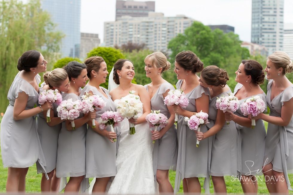 Bride with bridesmaids holding pink bouquets in a city park, smiling in gray dresses and white gown