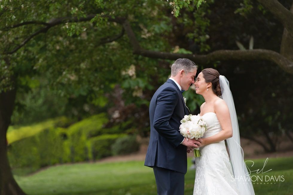 Bride and groom kissing outdoors under a leafy tree, bride holding a white bouquet.