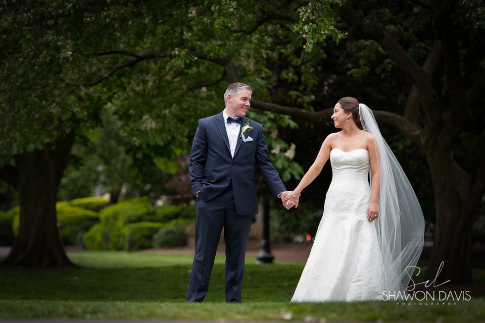 Bride and groom holding hands in a garden, smiling under a dark tree canopy