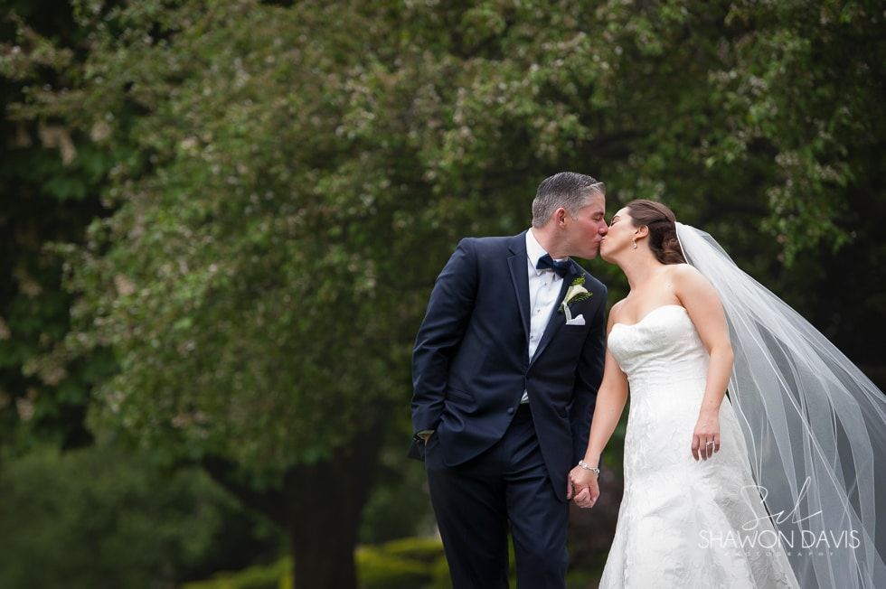Bride and groom kissing outdoors, with the bride’s veil flowing in the breeze