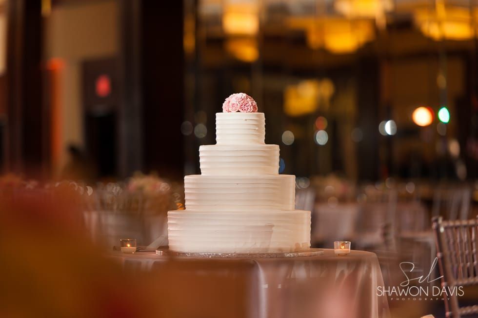Tiered white wedding cake with pink floral topper on a table in a warmly lit reception hall