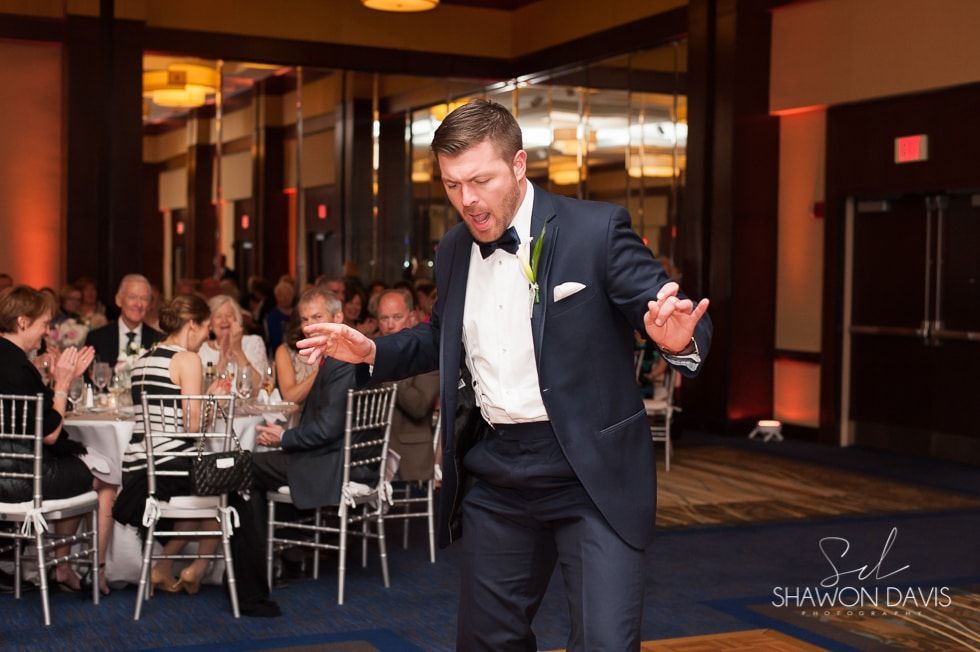 Man in a tuxedo dancing at a formal indoor reception with seated guests in the background