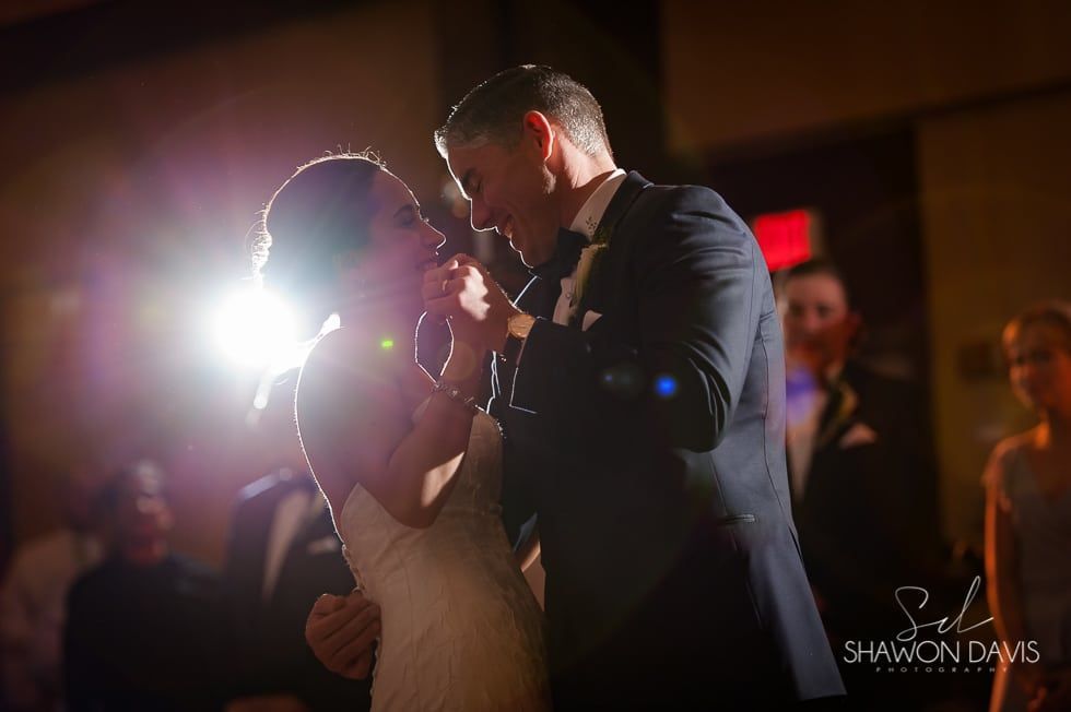 Couple slow dancing at a dimly lit wedding reception, backlit by bright stage lights