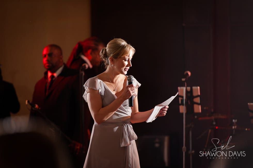 Woman in a white dress reading from a paper into a microphone on a dimly lit stage