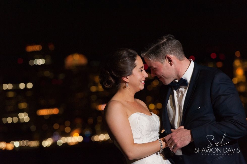 Bride and groom embracing at night with city lights in the background