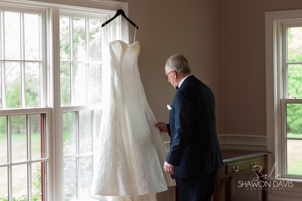 White wedding dress hanging by a bright window while a man in a suit looks at it