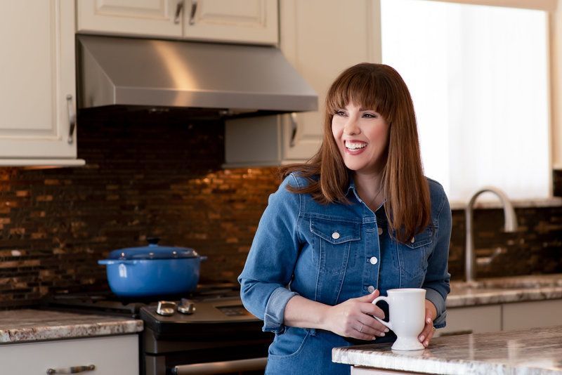Woman in a denim shirt smiling in a kitchen, holding a mug beside a stovetop and blue pot