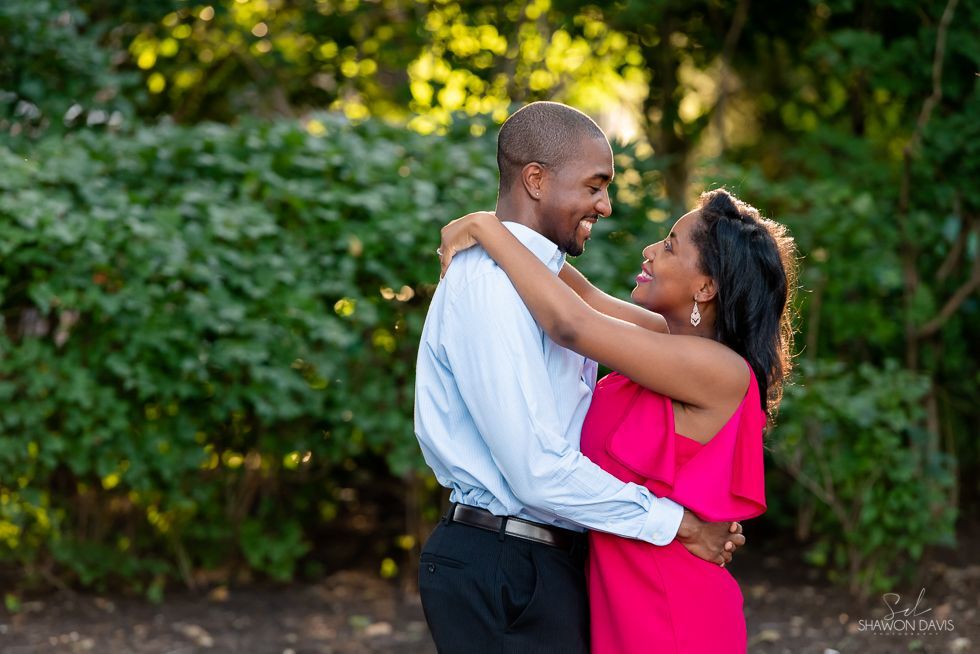 Couple embracing outdoors, smiling in a garden with green foliage and warm sunlight.