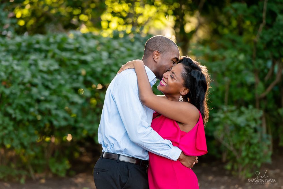 Two people embracing outdoors, smiling in a lush green garden; one wears a pink dress, the other a light blue shirt.
