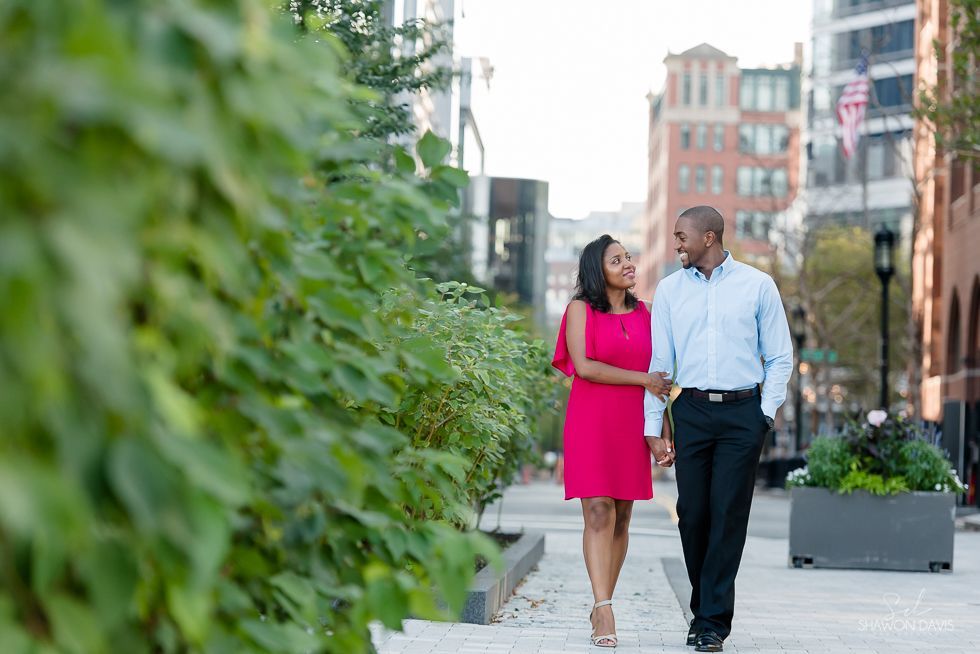 Two people walking and talking on a city sidewalk beside a green wall and potted plants.