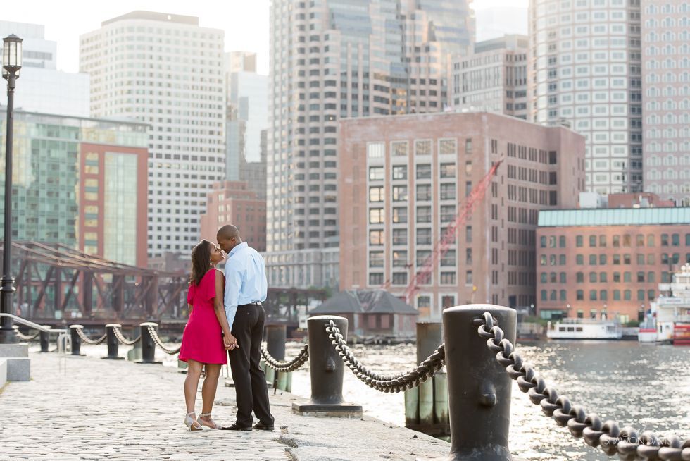 Couple standing by a waterfront railing with city skyline and bridge in the background