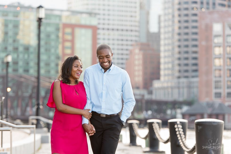 Two people walking and smiling on a city waterfront promenade, with high-rise buildings in the background.