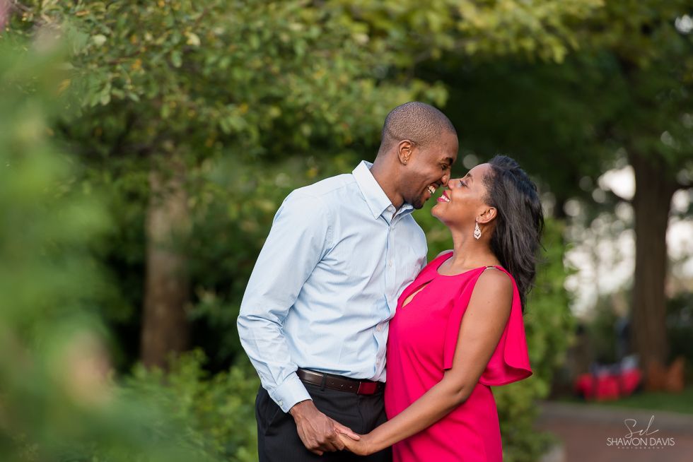 Couple kissing outdoors in a green park, with the woman in a bright pink dress and the man in a light blue shirt