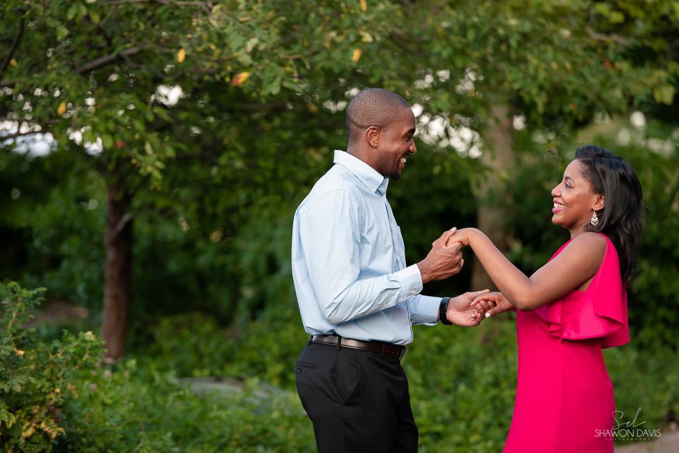 Two people smiling and holding hands in a green park, with one in a white shirt and the other in a pink dress
