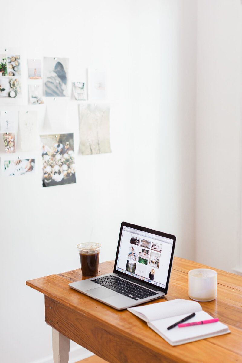 Minimalist desk with laptop, iced coffee, notebook, and candle beside a wall of pinned photos.