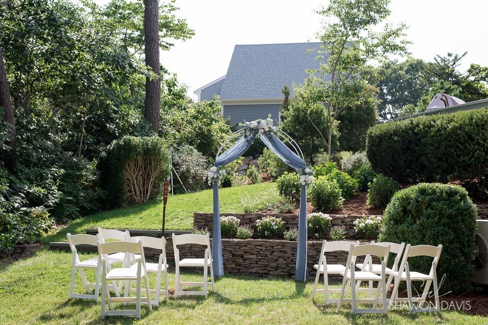White chairs under a wedding arch on a lawn in a garden with a house in the background