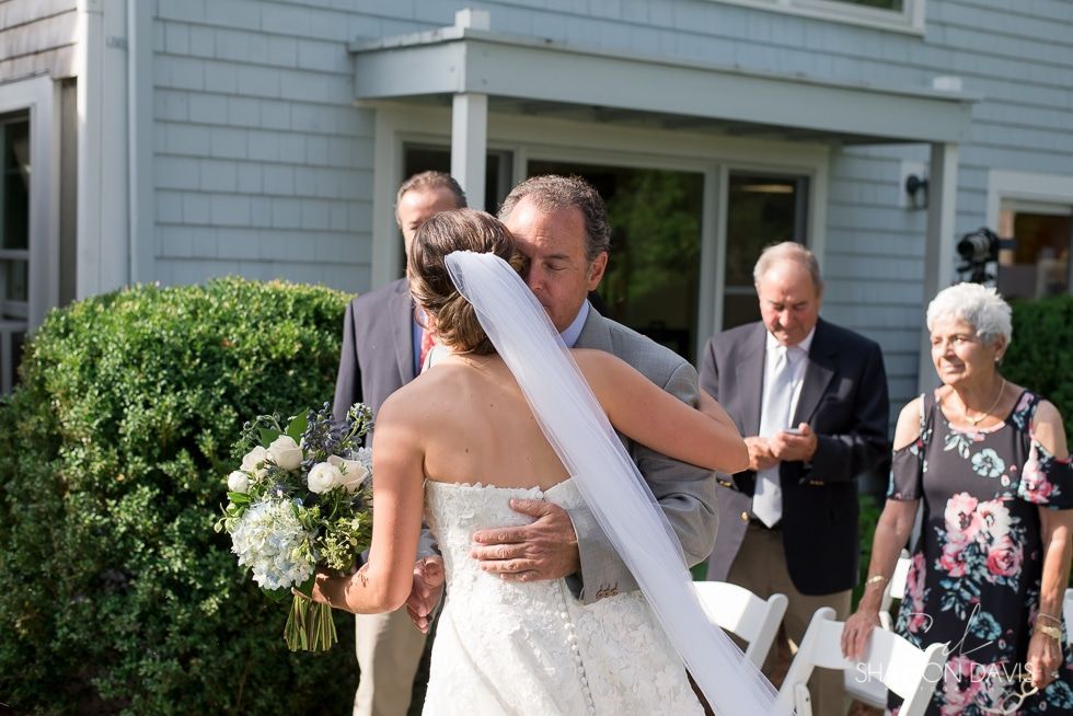 Bride in a white dress hugs a man outdoors at a wedding, with guests and a house in the background.