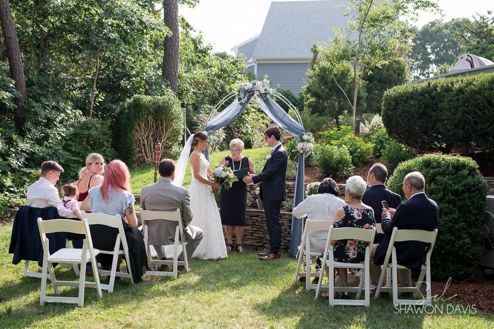 Outdoor wedding ceremony under a floral arch, guests seated on white chairs in a sunny garden