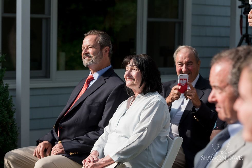 Seated audience members outdoors in sunlight, one man reading a red phone, others in formal attire.