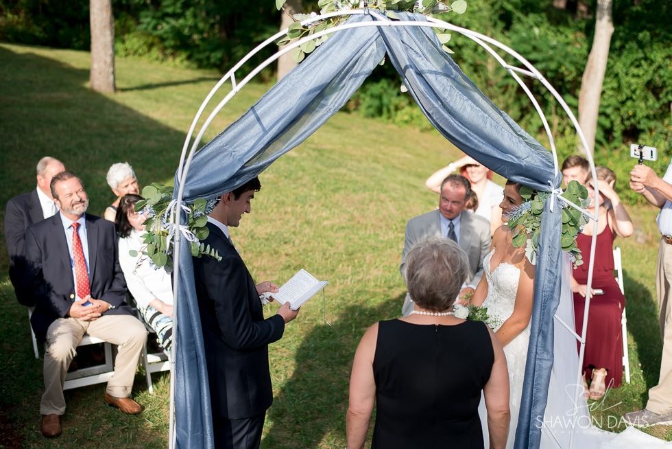 Outdoor wedding ceremony under a blue-and-white arch, with guests seated and the bride and groom at the altar