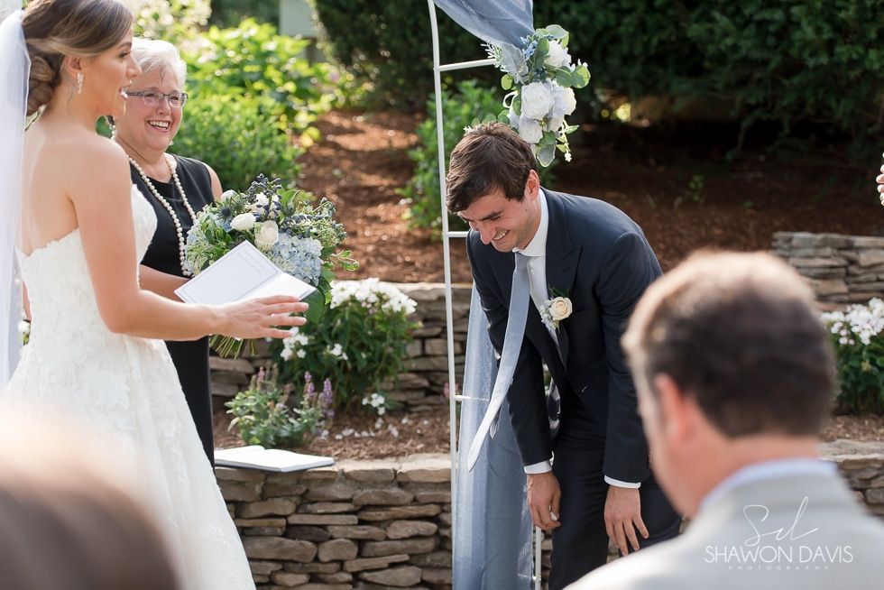 Outdoor wedding ceremony with bride reading vows to groom under a floral arch by a stone wall