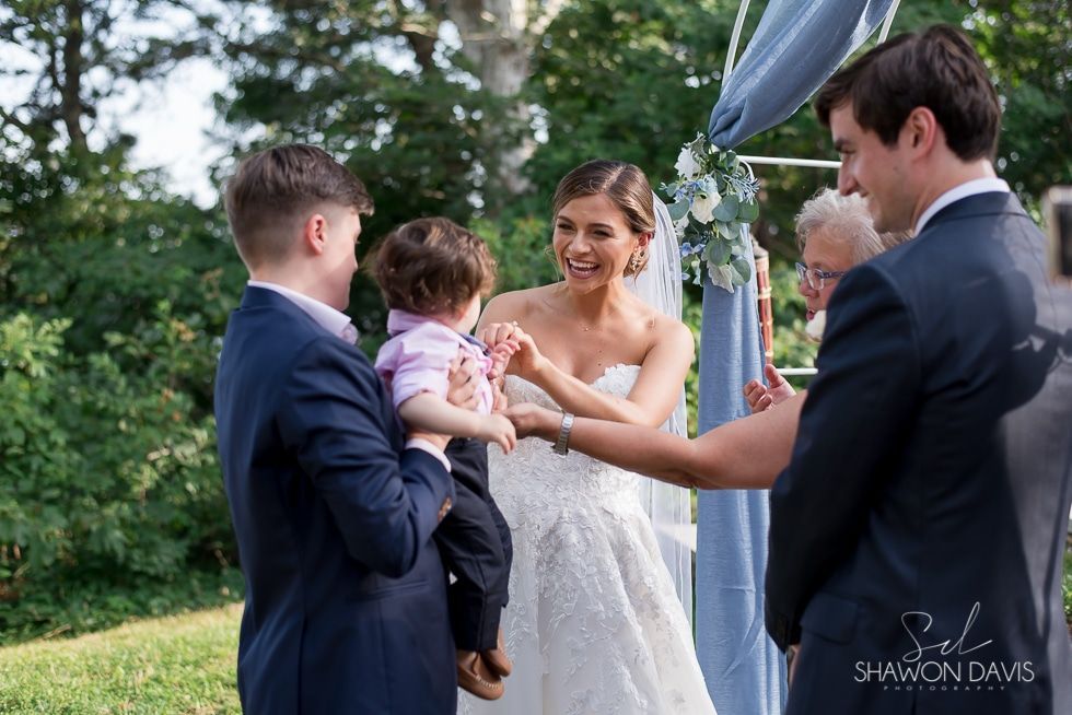 Bride smiling at a wedding ceremony, reaching toward a child held by a man under a canopy outdoors