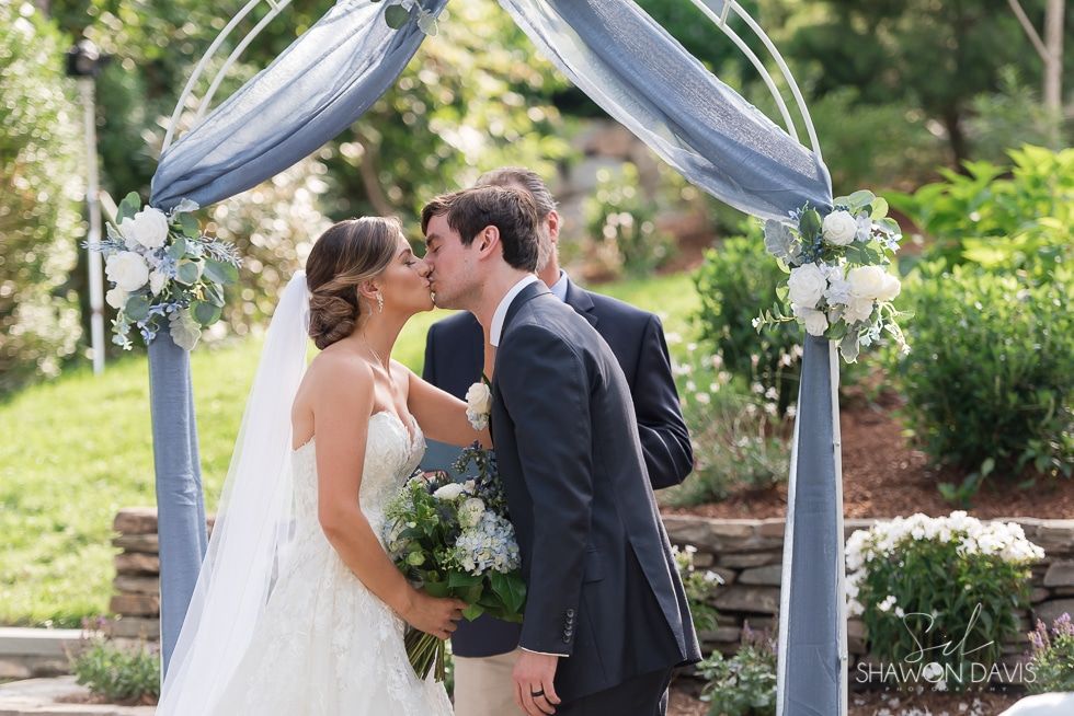Bride and groom kissing under a decorated outdoor wedding arch in a garden