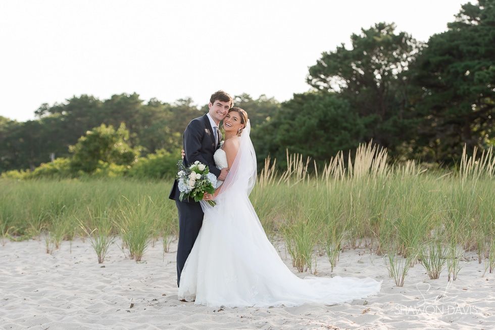 Bride and groom embracing on a sandy beach beside tall grass at sunset