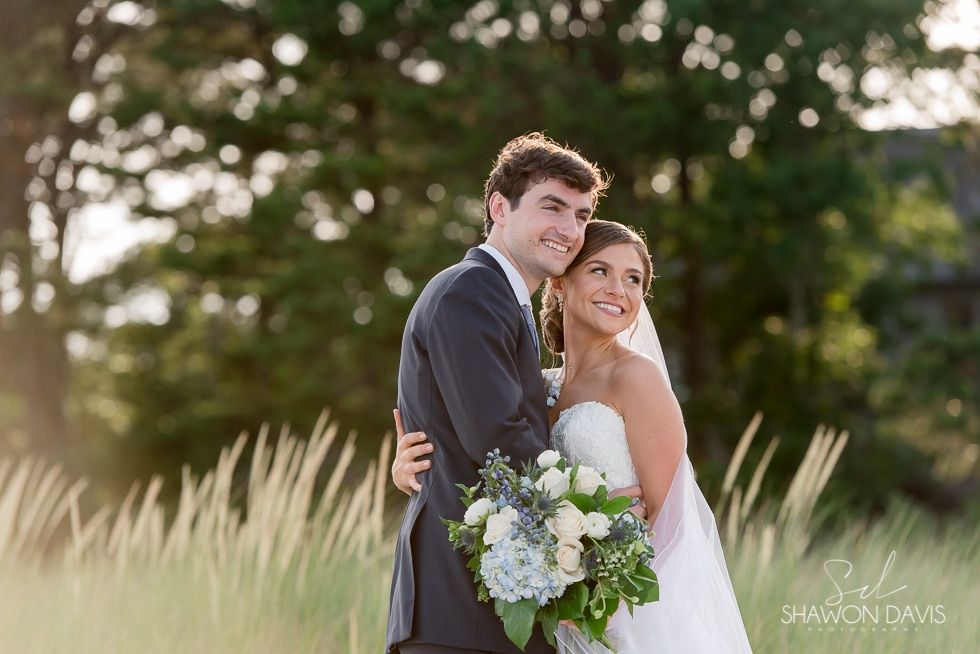 Bride and groom smiling in a field, holding a bouquet during an outdoor wedding ceremony.