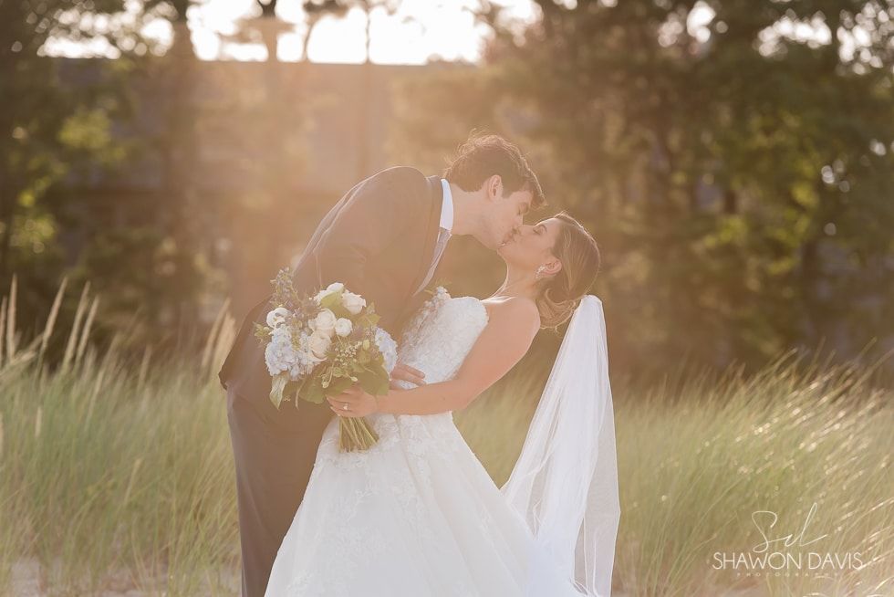 Bride and groom kiss in a sunlit field, with warm golden light and blurred trees behind them