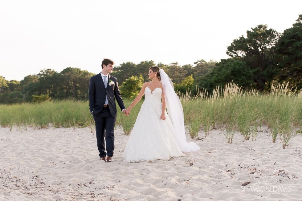Bride and groom holding hands on a sandy beach with grasses and trees in the background