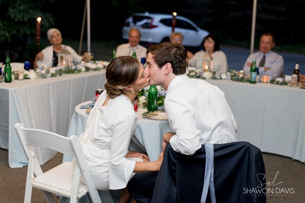 Two people in white shirts kiss at a candlelit reception table with seated guests nearby.