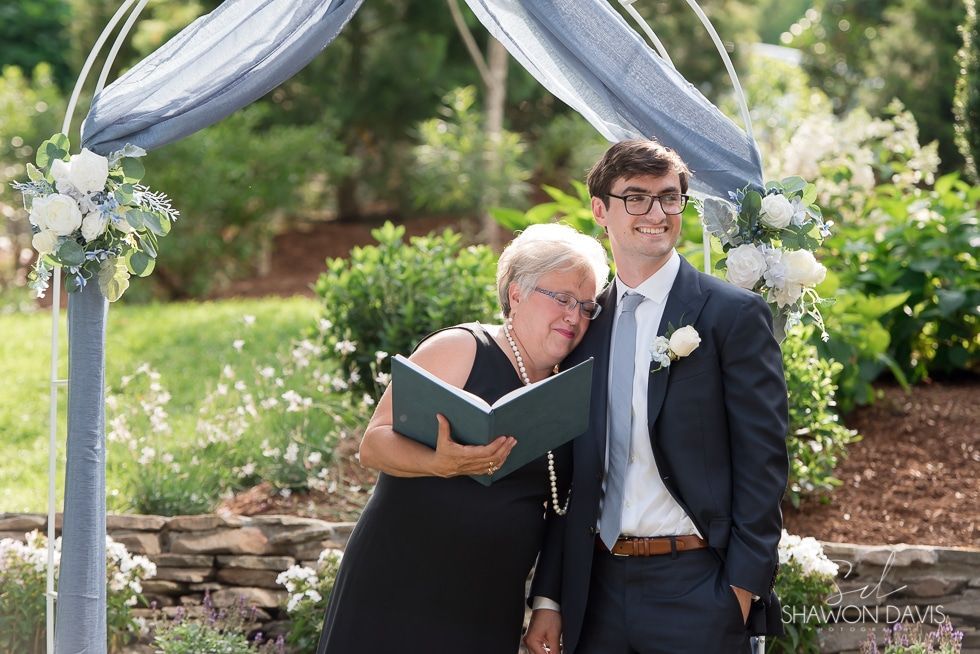 Two people in formal wear standing under a floral wedding arch outdoors, reading from a green book