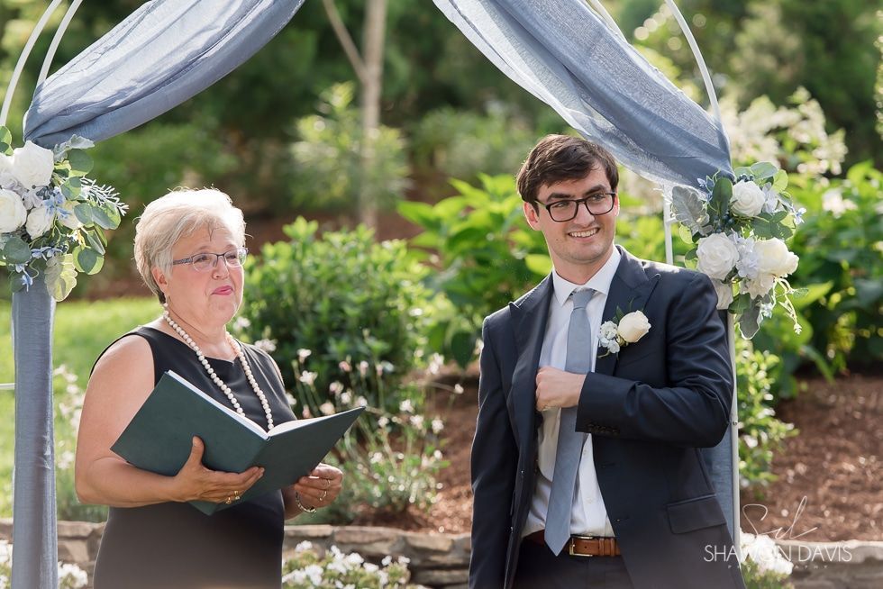 Two people standing under a blue and white canopy during an outdoor ceremony, one reading from a book.
