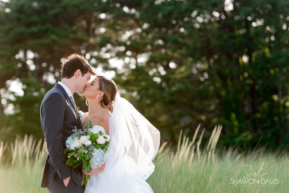 Wedding couple kissing outdoors in a grassy field, with trees in the background.