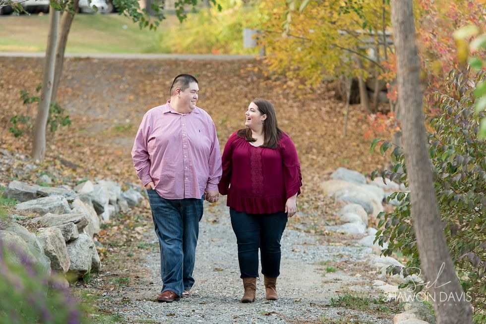 Two people walking hand in hand on a leaf-strewn path in a wooded park, smiling at each other.