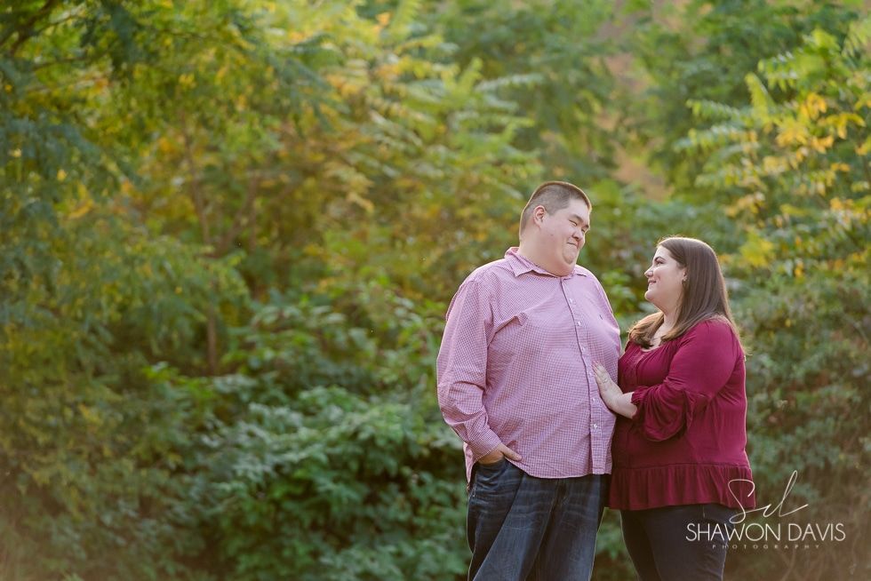 Couple standing together in a wooded outdoor setting, looking at each other and smiling.