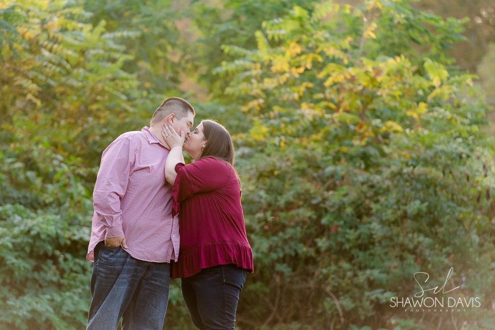 Two people kissing outdoors in front of green foliage; one wears a pink shirt, the other a red sweater.