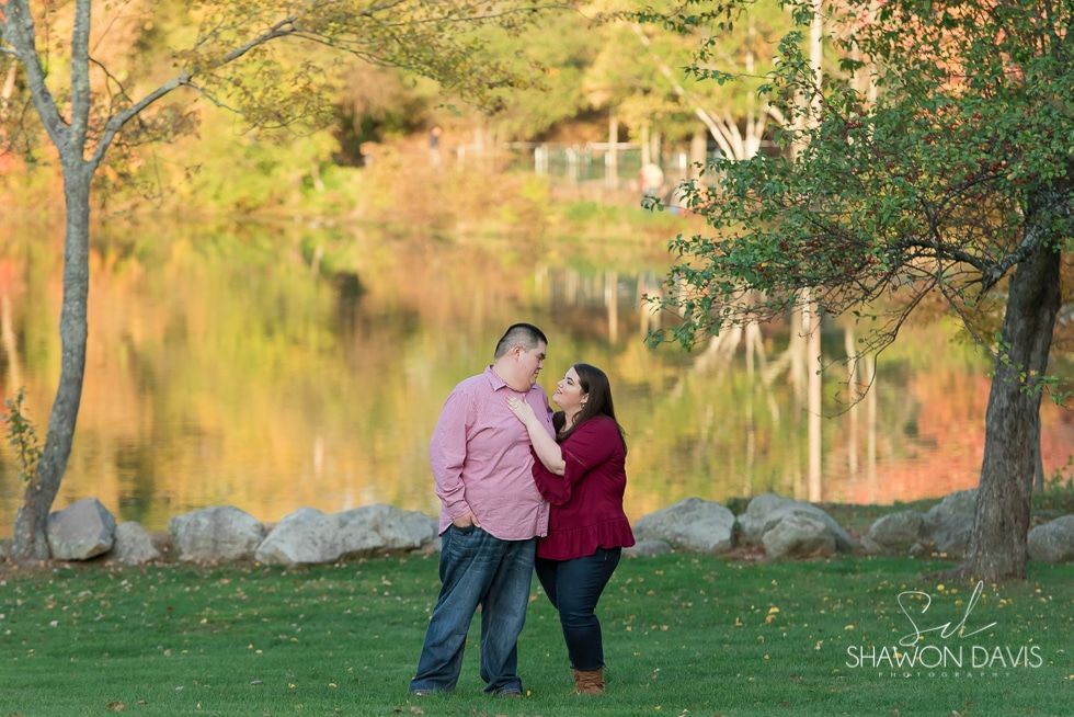 Couple standing close in a sunlit park by a lake with autumn trees and rocks in the background