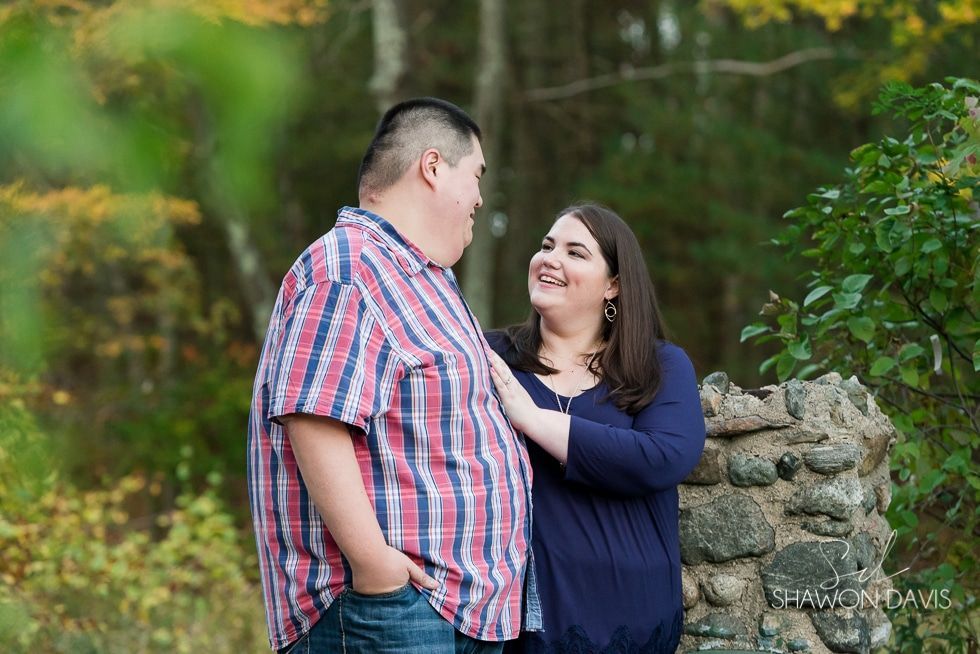 Two people smiling outdoors, standing close beside a stone wall with greenery behind them