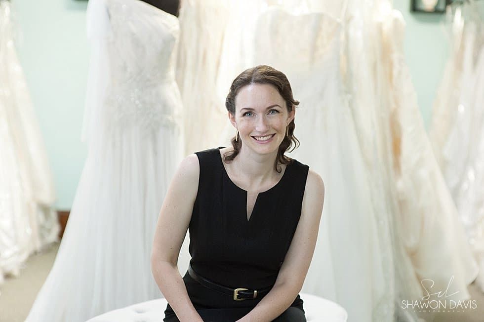 Smiling woman in a black sleeveless dress seated in a bridal shop, with white gowns in the background