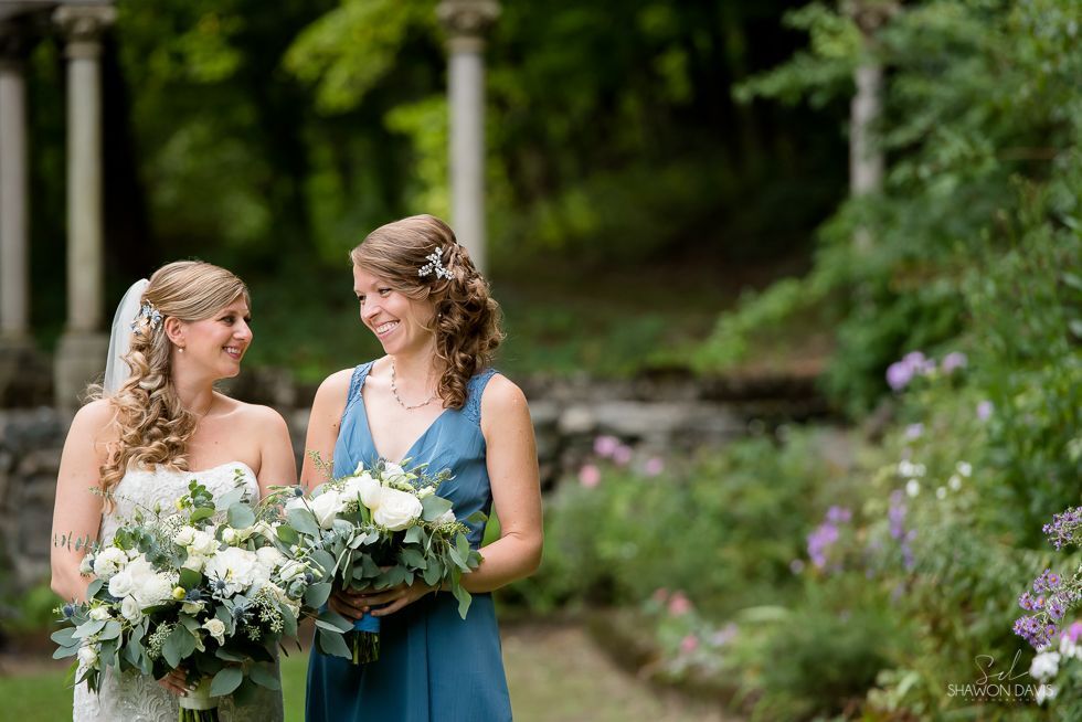 Two women in blue dresses holding bouquets outdoors in a garden, smiling at each other