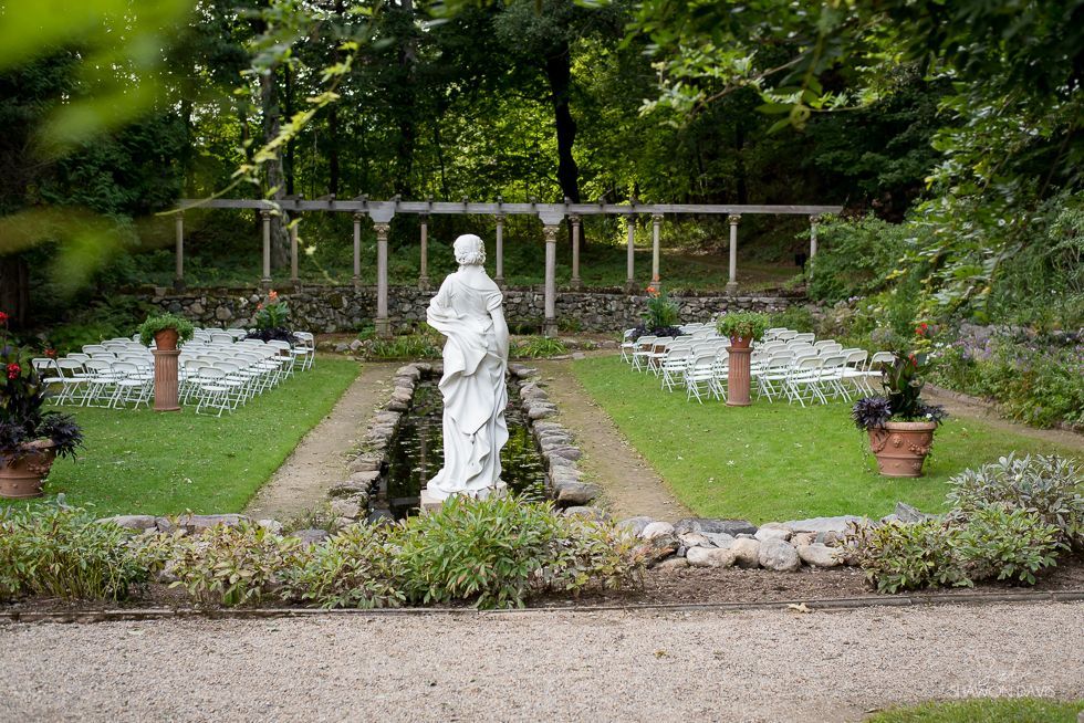 White statue in a formal garden path surrounded by flowers and greenery, with a wooden trellis in back