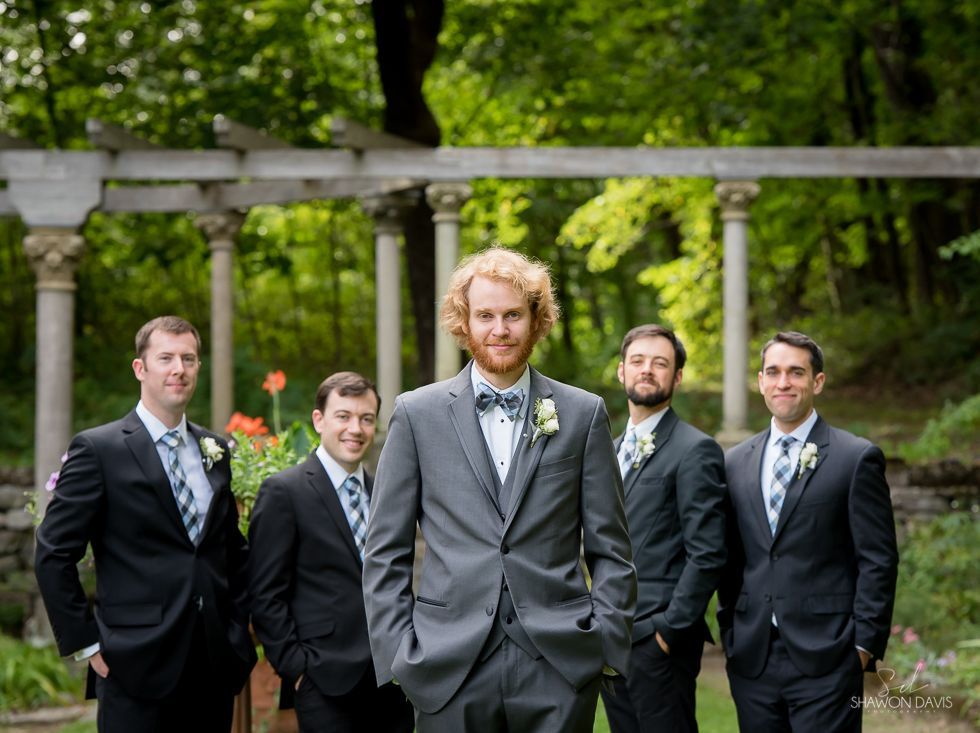 Five men in suits posing outdoors in a garden with stone columns and greenery.