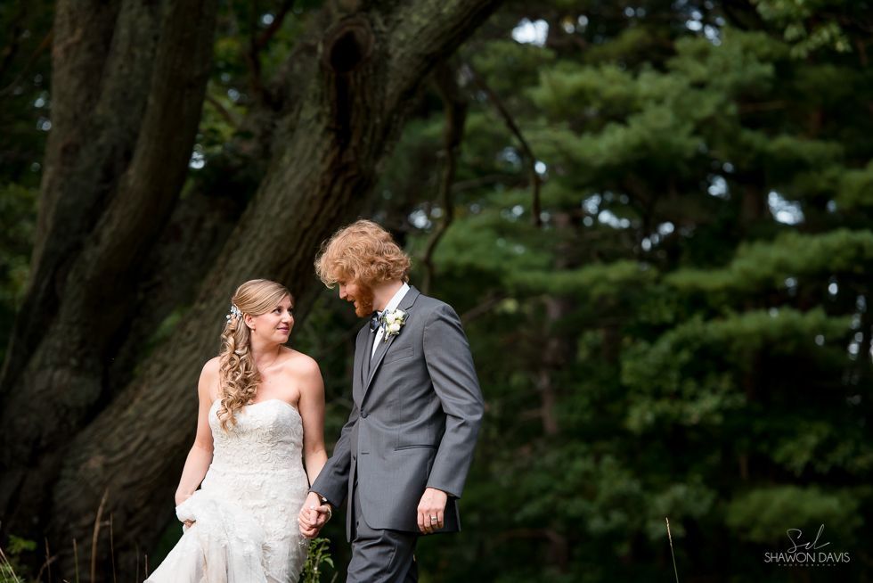 Bride and groom smiling in a garden, standing close beneath large trees.