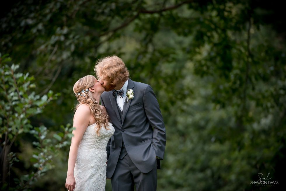 Bride and groom kissing outdoors in a lush green garden, the bride in a white lace dress and the groom in a dark suit.