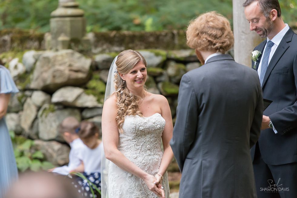Bride smiling at groom during outdoor wedding ceremony with guests and stone garden backdrop